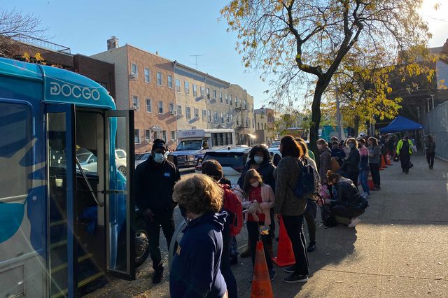 Students and parents on a sidewalk, waiting outside a vaccine bus, on a sunny day